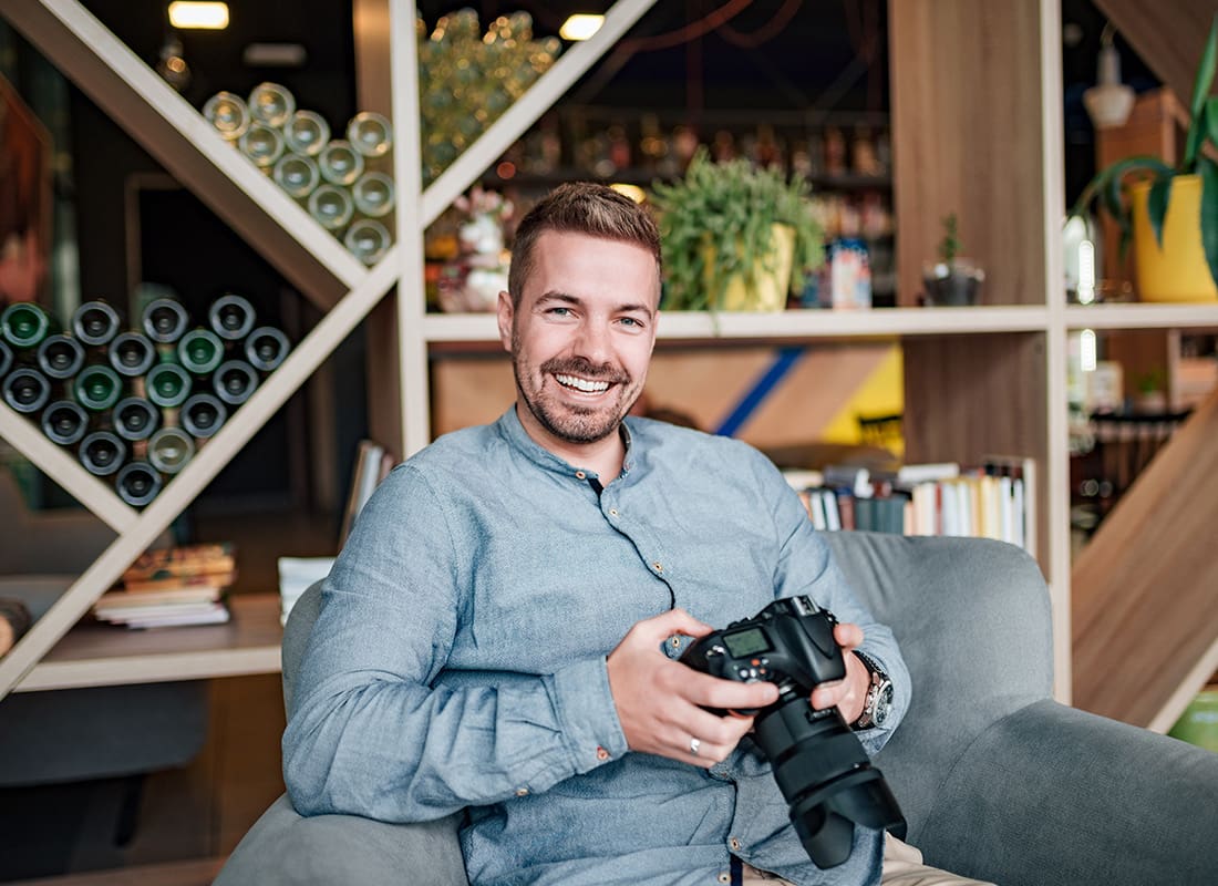 Business Insurance - Cheerful Man Holding a Camera While Sitting Down in His Studio